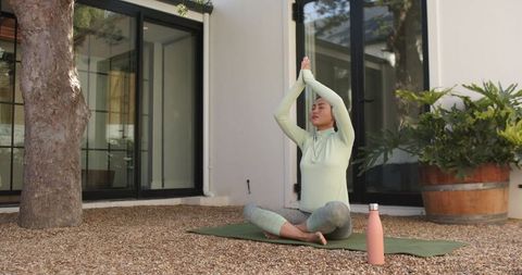 Asian woman practicing outdoor yoga on pebble patio with plants and pink water bottle