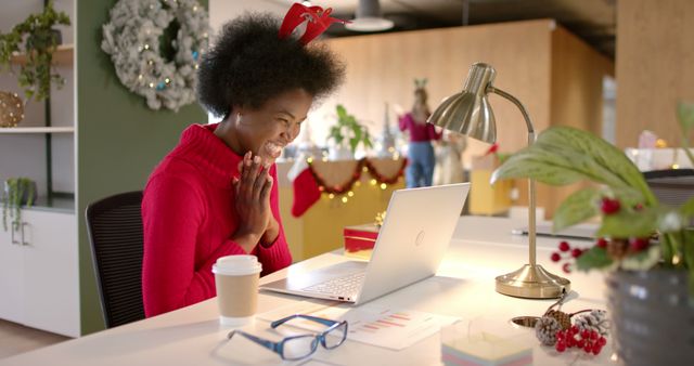 Woman in festive office environment smiling at laptop while participating in a virtual holiday party. Ideal for themes related to Christmas celebrations, remote work interactions, virtual events, holiday festivities, and workplace holiday spirit.