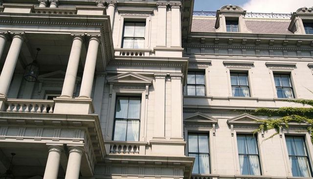 Sunlit neoclassical stone facade featuring fluted columns, pediments, balustrade and ivy