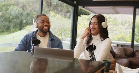 Diverse Cohosts Laughing and Recording Podcast in Bright Sunroom with Laptop and Mics