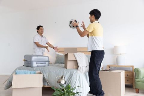 Asian men unpacking boxes in bright minimalist bedroom passing soccer ball during move