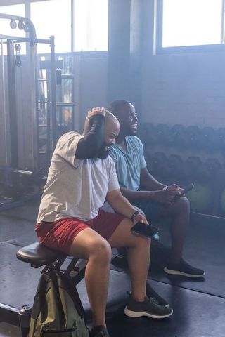 African male friends laughing during workout break on gym bench holding smartphones