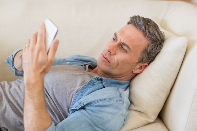 Man lying on sofa and using mobile phone in living room at home