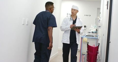 Asian doctor and black nurse consulting tablet in hospital corridor with wheelchair