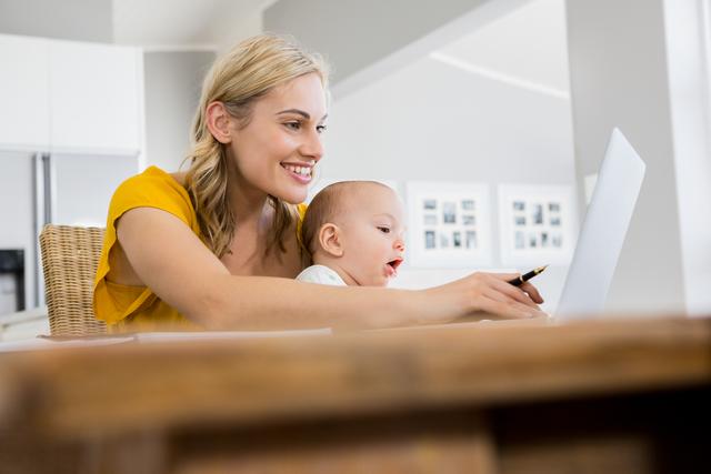Depicts a mother multitasking by using a laptop while holding her baby boy in a home kitchen. Perfect for illustrating modern parenting, work-from-home scenarios, and family life. useful for articles or advertisements on work-life balance, remote work, technology in daily life, and positive family moments.