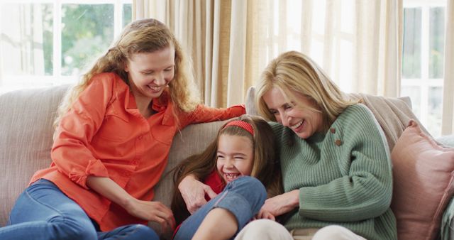 Three women from different generations, including a grandmother, mother, and young daughter, are relaxing and bonding on a couch. They are visibly happy and engaged, enjoying quality family time together. This heartwarming scene is ideal for illustrating concepts of family, love, multigenerational relationships, and togetherness. It can be used for family-oriented campaigns, articles on family dynamics, or advertisements for household products that promote spending time together.