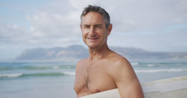 Portrait of happy shirtless senior caucasian man holding surfboard on sunny beach. Sport, surfing, hobbies, vacations and outdoor activities, unaltered.