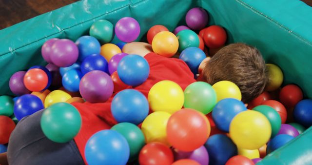 Child lying in a ball pit filled with multicolored balls at an indoor playground. Perfect for use in advertisements for family-friendly venues, articles on child development and play, or marketing materials for indoor recreational facilities.