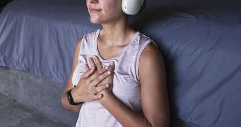 Young woman meditating with headphones, hands over heart, practicing breathwork near bed