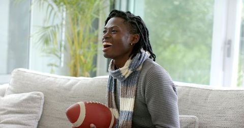African American man cheering on sofa holding football wearing cozy scarf and sweater