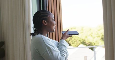 African american woman speaking into smartphone by balcony smiling and relaxing at home