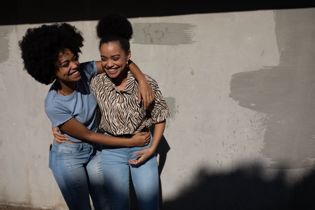 Front view of two happy biracial twin sisters enjoying free time together, embracing, smiling, standing by a wall on a city street on a sunny day.