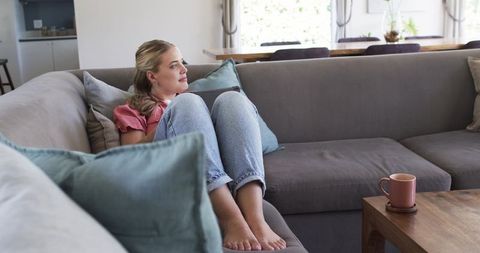 Young woman curling on grey sofa gazing through window cozy living room neutral tones