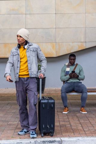 Young traveler standing with suitcase while friend checking phone at urban transit stop