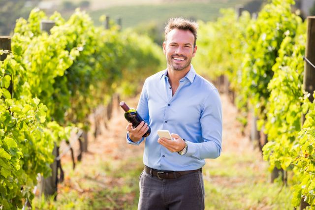 Man standing in vineyard holding wine bottle and using phone, smiling on sunny day. Ideal for use in advertisements for wineries, wine tours, agricultural technology, rural lifestyle promotions, and business casual attire. Highlights combination of traditional viticulture with modern technology.