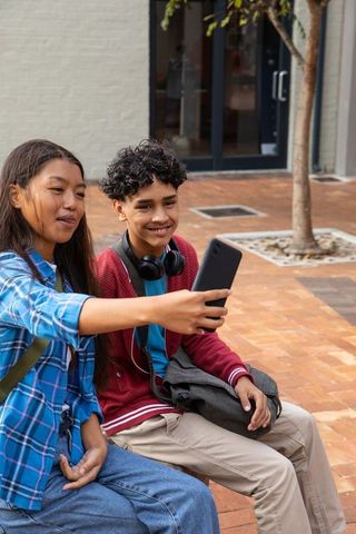 Asian teen girl and boy taking selfie on urban bench with smartphone and headphones