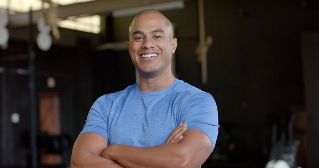 Portrait of happy biracial man wearing blue tshirt with arms crossed at gym. Activity, sport and exercise, unaltered.