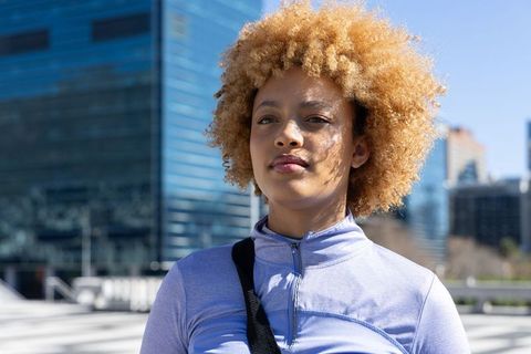 African american woman standing in sunlit city plaza wearing athletic zip-up top and bag