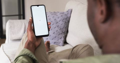 African american man reclining on beige sofa holding smartphone with blank screen