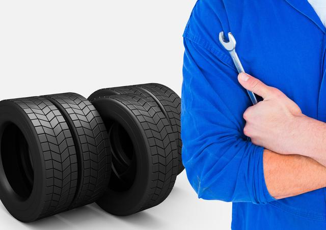 Mechanic wearing blue uniform holding wrench with two tires in background. Useful for illustrating auto repair, tire service, and car maintenance themes.