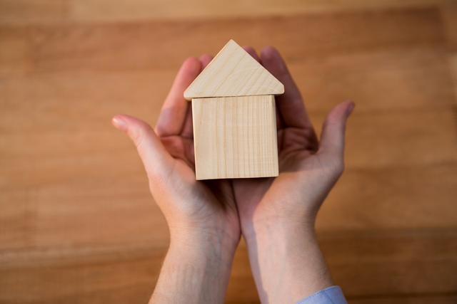 Close-up view of hands gently holding a wooden miniature house, symbolizing real estate, homeownership, and security. Ideal for use in real estate advertisements, home insurance promotions, investment brochures, and articles about housing and property care.