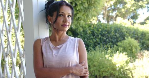 Sunlit contemplative woman leaning on porch post, crossing arms and gazing into garden