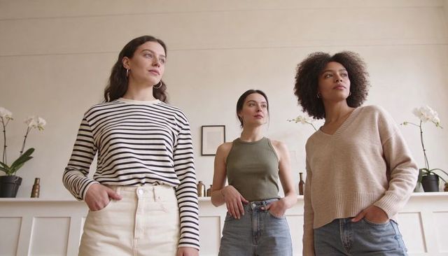 Three women standing in stylish living room wearing casual denim and neutral knitwear