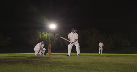 Night cricket under floodlights with batsman batting, wicketkeeper crouching, outfielder waiting