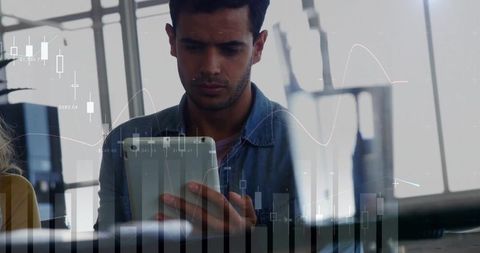 Holding tablet studying financial charts in backlit office with colleague