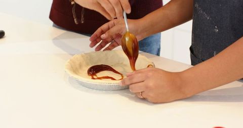 Couple filling pie crust with caramel in bright kitchen, spoon drizzling filling