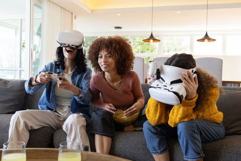 Diverse women playing virtual reality games and sharing popcorn on cozy living room sofa
