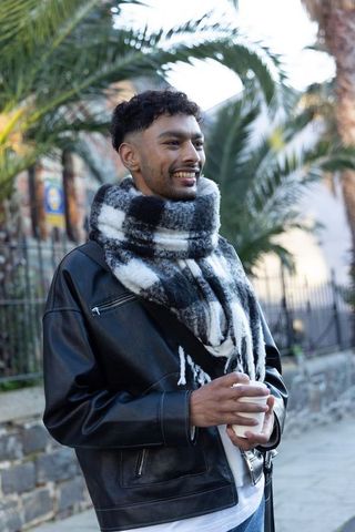 Smiling young man holding takeaway coffee cup wearing leather jacket and checked scarf