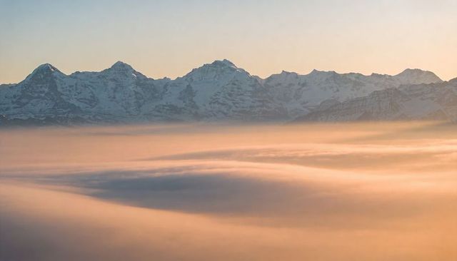 Snow-covered mountain range glowing above sea of clouds at sunrise, soft pastel dawn light