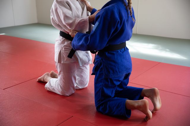 Low section of two Caucasian female judokas wearing blue and white judogi, practicing judo on a mat during a training in a bright studio.