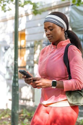 African woman checking phone on urban sidewalk wearing headband, smartwatch, workout gear