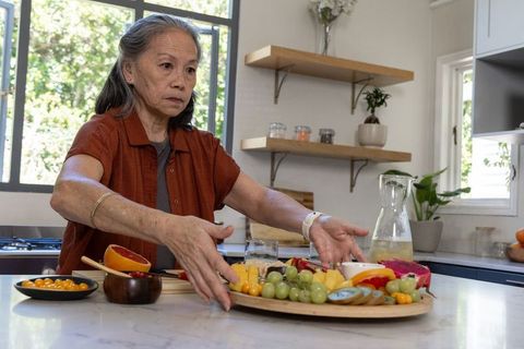 Mature asian woman arranging colorful gourmet fruit platter on marble kitchen island