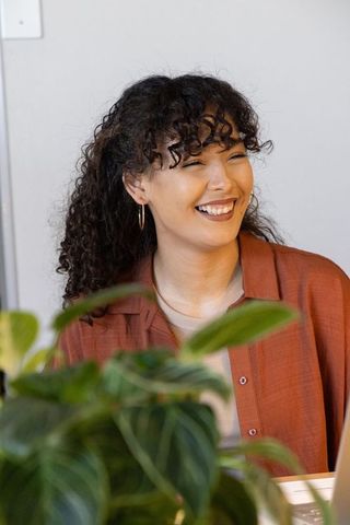 Young woman smiling while listening at home desk with laptop and lush indoor plant