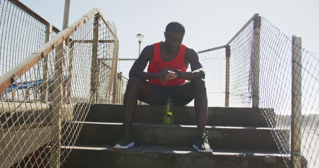 Young black man sitting on outdoor stairs, watching smartwatch after intense workout. Wearing red tank top and athletic shoes. Scene highlights fitness, determination, and exhaustion. Ideal for use in articles about fitness routines, smartwatches in sports, and training advertisements.
