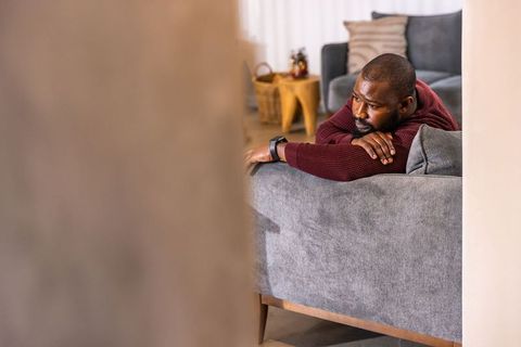 Contemplative african american man reclining on gray sofa wearing smartwatch at home