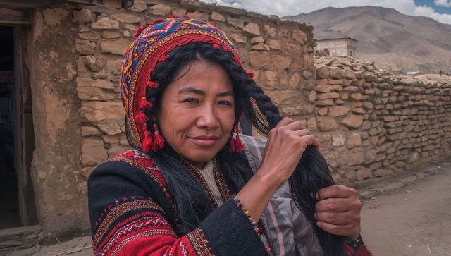 Andean woman adjusting braided hair in colorful woven hat and embroidered jacket, adobe village