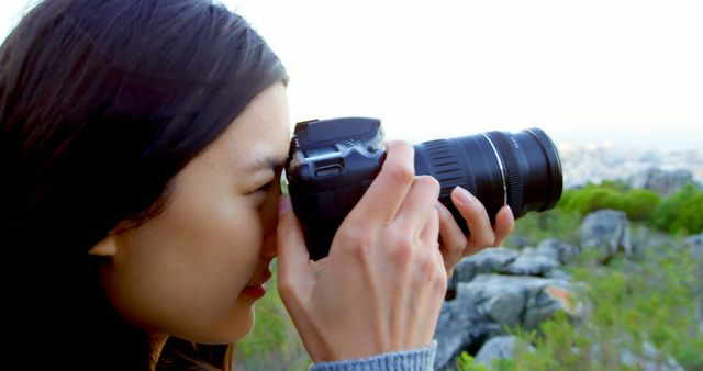 Young woman holding a DSLR camera focused on photographing nature. Background features rocky terrain and green foliage. Ideal for topics related to hobbies, outdoor activities, photography techniques, and creative pursuits.