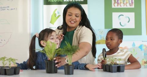 Diverse female teacher guiding students examining seedlings with magnifying glass during classroom s