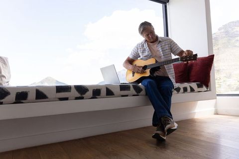 Man playing acoustic guitar on window bench with laptop in minimalist modern interior