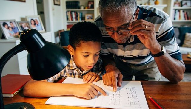 Grandfather guiding grandson through math workbook under desk lamp, family learning moment