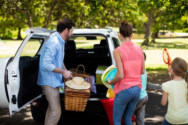 Family packing picnic items into car trunk at park. Parents and children preparing for a day out, placing baskets and blankets into the vehicle. Ideal for concepts related to family bonding, outdoor activities, summer fun, and weekend getaways.
