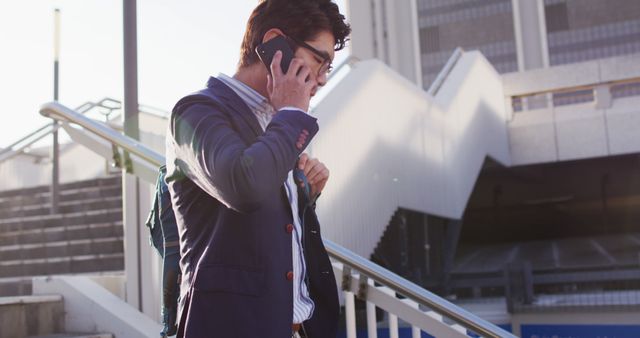 Asian businessman wearing suit talking on smartphone outdoors in an urban city setting. He appears professional and in a rush, holding a shoulder bag. This image can be used for themes related to business, urban lifestyle, communication technology, and professional attire.