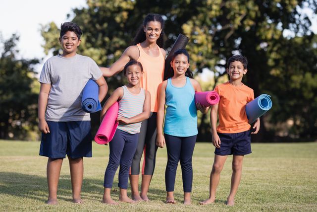 Group of children with female instructor holding yoga mats in a park, smiling and ready for a fitness session. Ideal for promoting outdoor activities, family bonding, healthy lifestyle, and fitness programs for kids.
