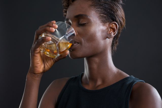 Androgynous man enjoying a glass of whiskey against a dark background. Ideal for use in lifestyle blogs, articles on relaxation and leisure, or advertisements for alcoholic beverages. The image conveys a sense of calm and enjoyment, perfect for marketing campaigns or editorial content focused on personal time and indulgence.