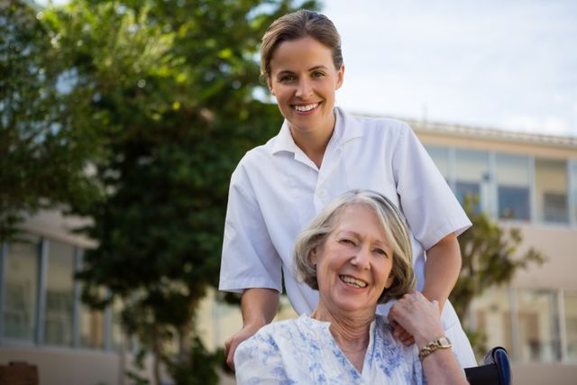 This image depicts a cheerful caregiver standing behind a happy senior woman sitting in a wheelchair, both smiling. The outdoor setting suggests a pleasant environment, possibly a nursing home or retirement community. This image is ideal for use in healthcare, elderly care, and retirement home promotional materials, illustrating compassionate care and support for the elderly.