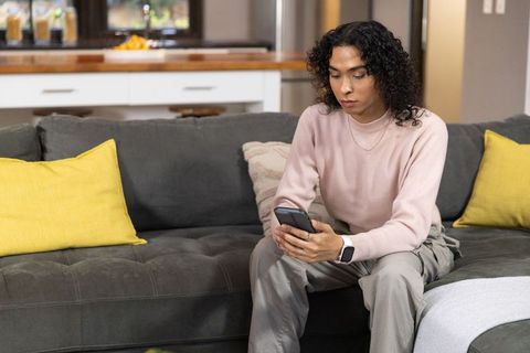 Nonbinary person on gray sofa scrolling smartphone, wearing smartwatch and pink sweater